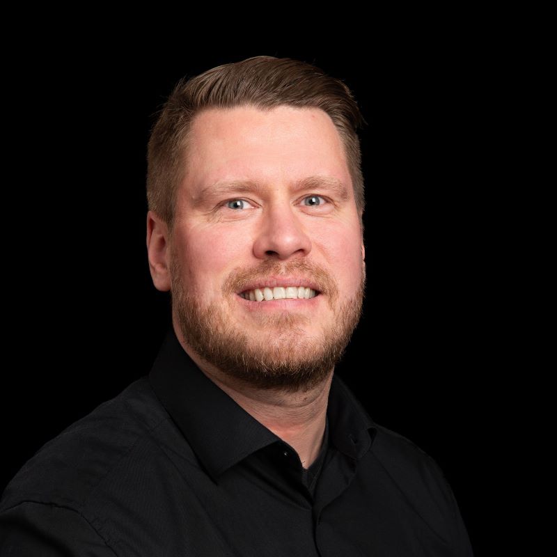 Hartioihin asti kuvattu mies mustassa paidassa tummalla taustalla.