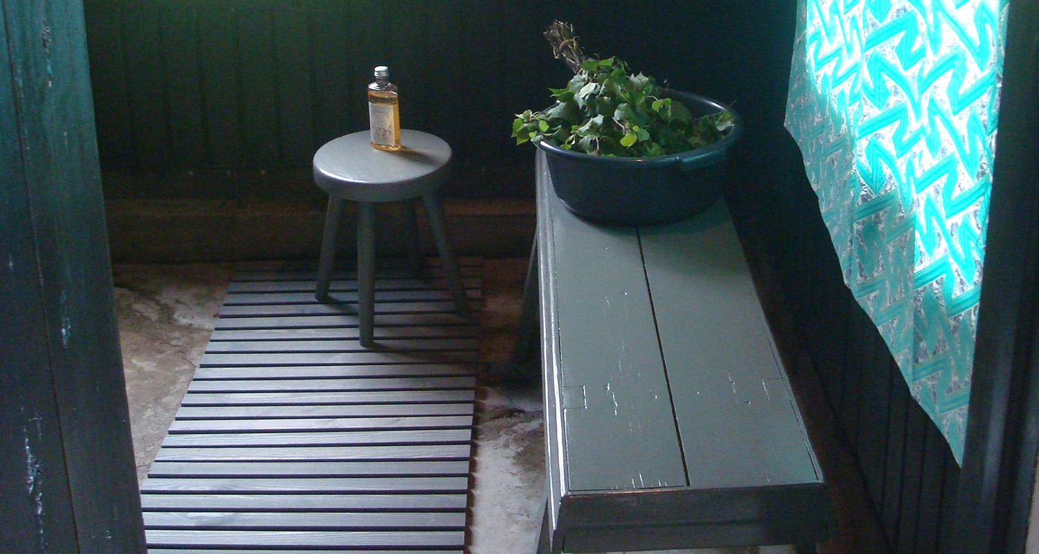 Saunan tummasävyinen eteinen: jakkaralla öljypullo, penkillä ämpärissä koivuvitsoja.