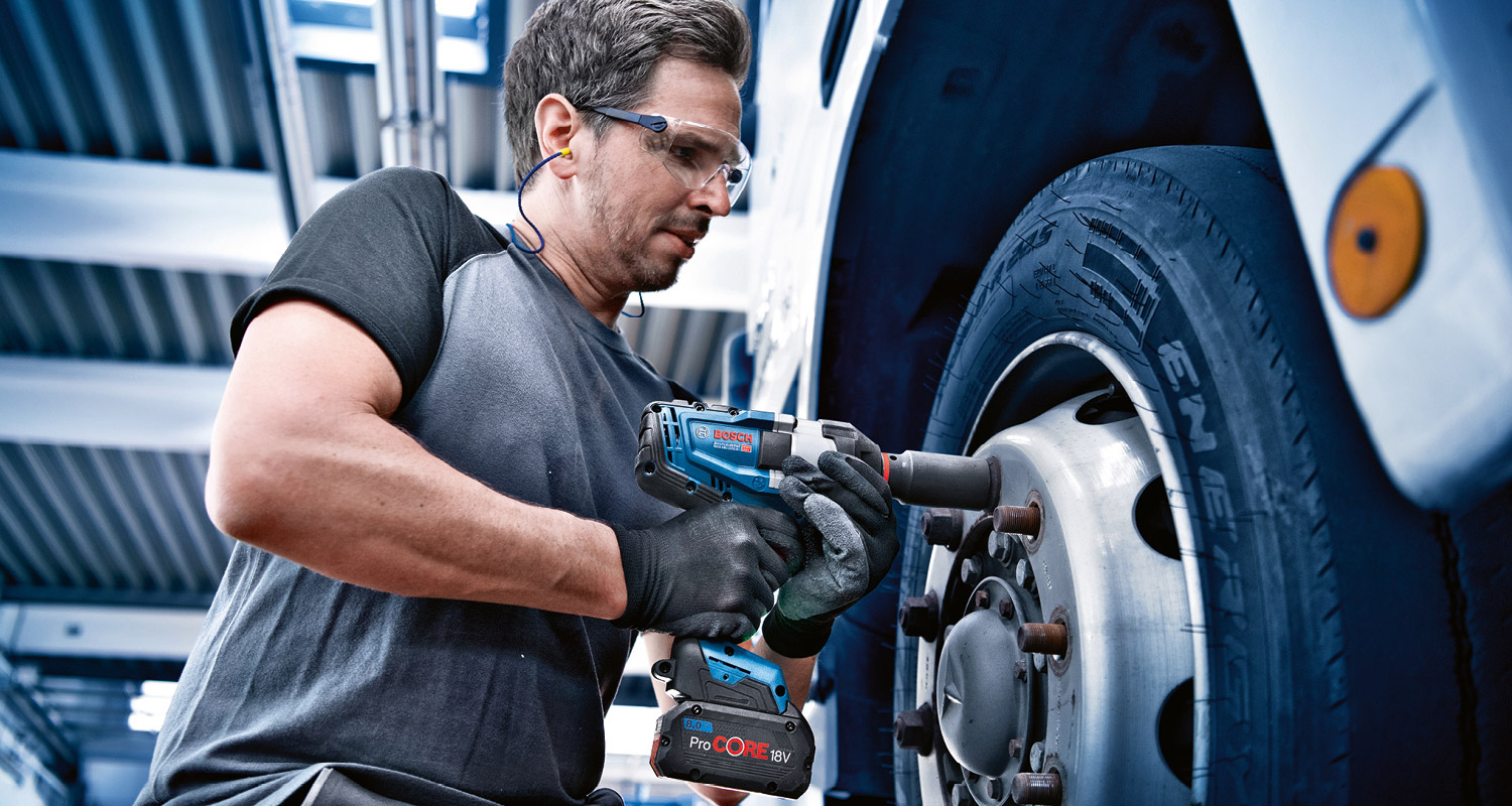 Harmaapaitainen asentaja käyttää sinimustaa 18V-iskuavainta kuorma-auton renkaan pulttiin.