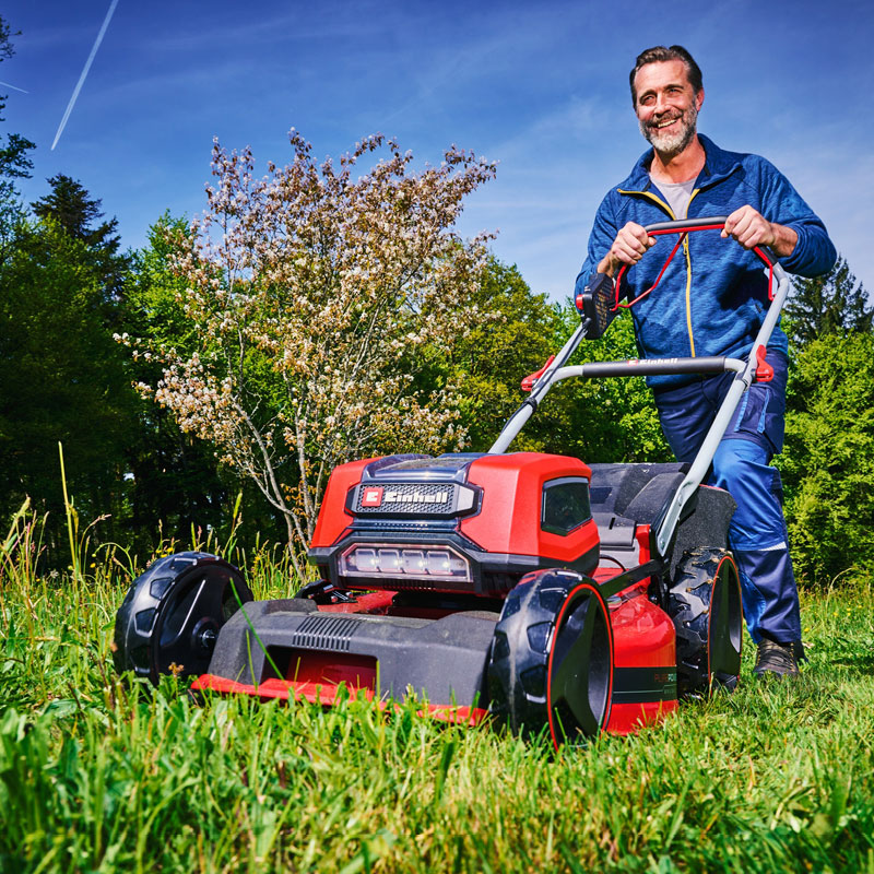 Mies työntää punaista ruohonleikkuria vihreällä nurmikolla aurinkoisessa puutarhassa.