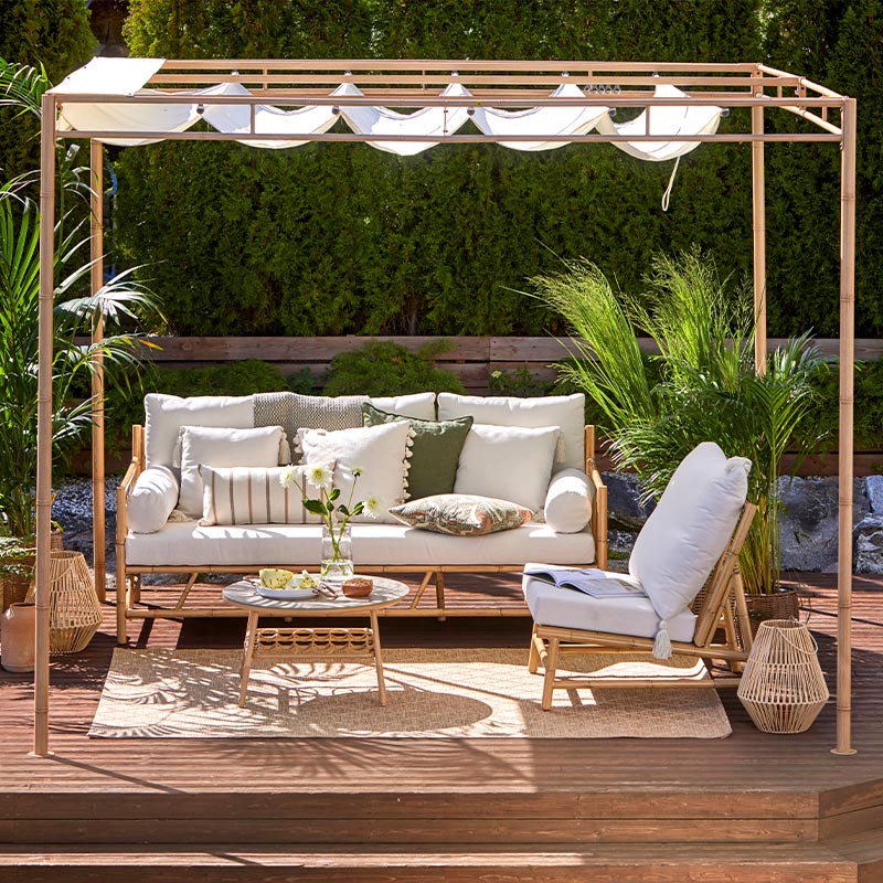 Terassilla pergola valkoisella kankaalla, valkoinen sohva ja nojatuoli tyynyin, viherkasveja.