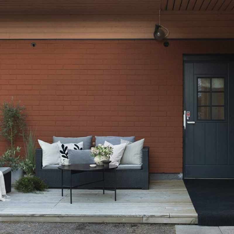 Punatiilinen seinä terassilla, musta ovi, harmaa ulkosohva valkoisilla tyynyillä ja kukkapöytä.