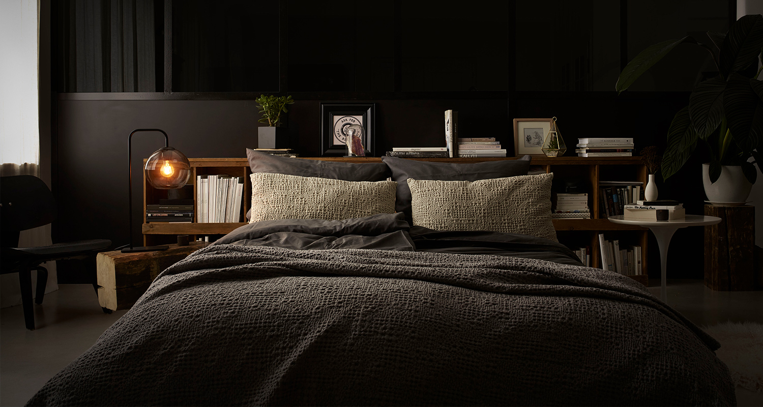 Tumma makuuhuone, harmaa peitto ja vaalea tyyny, päätyhyllyssä lamppu ja kirjapinot.