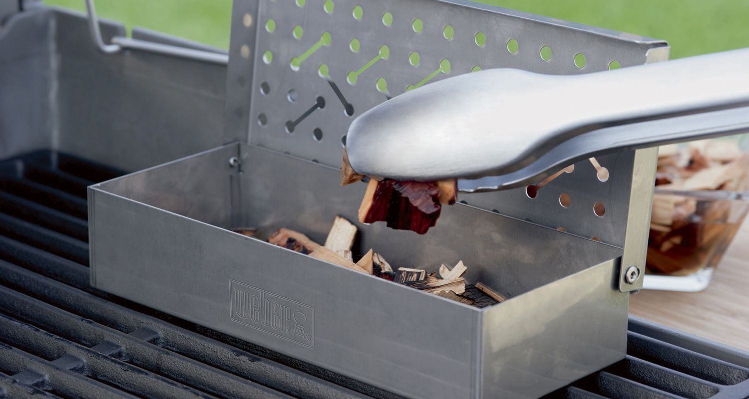Harmaa Weber-savustuskotelo grilliritilällä, metallitangolla lisätään ruskeita savupuruja.