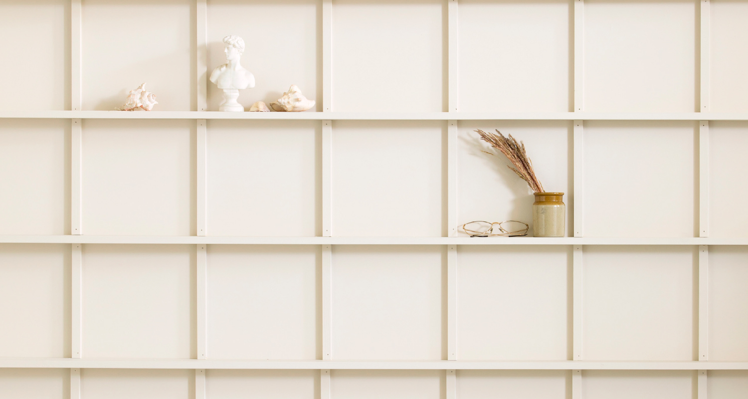 Valkoinen ruutuhylly, jossa klassinen rintakuva, simpukoita, purkissa kuivat tähkät ja silmälasit.