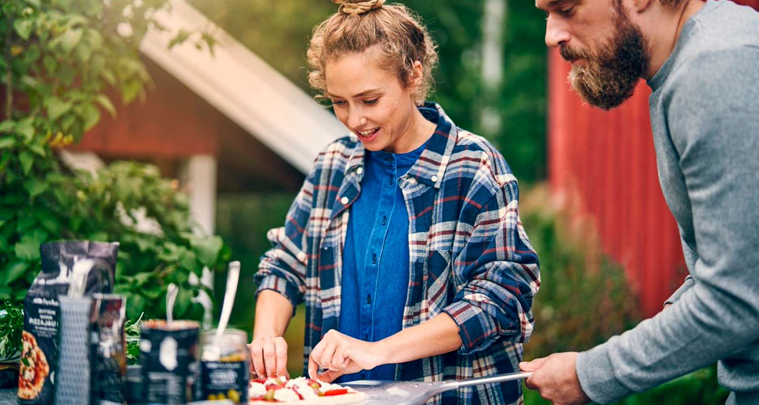 Ulkona ruutupaitainen muovaa pizzapohjaa, toinen ojentaa pizzalapion.