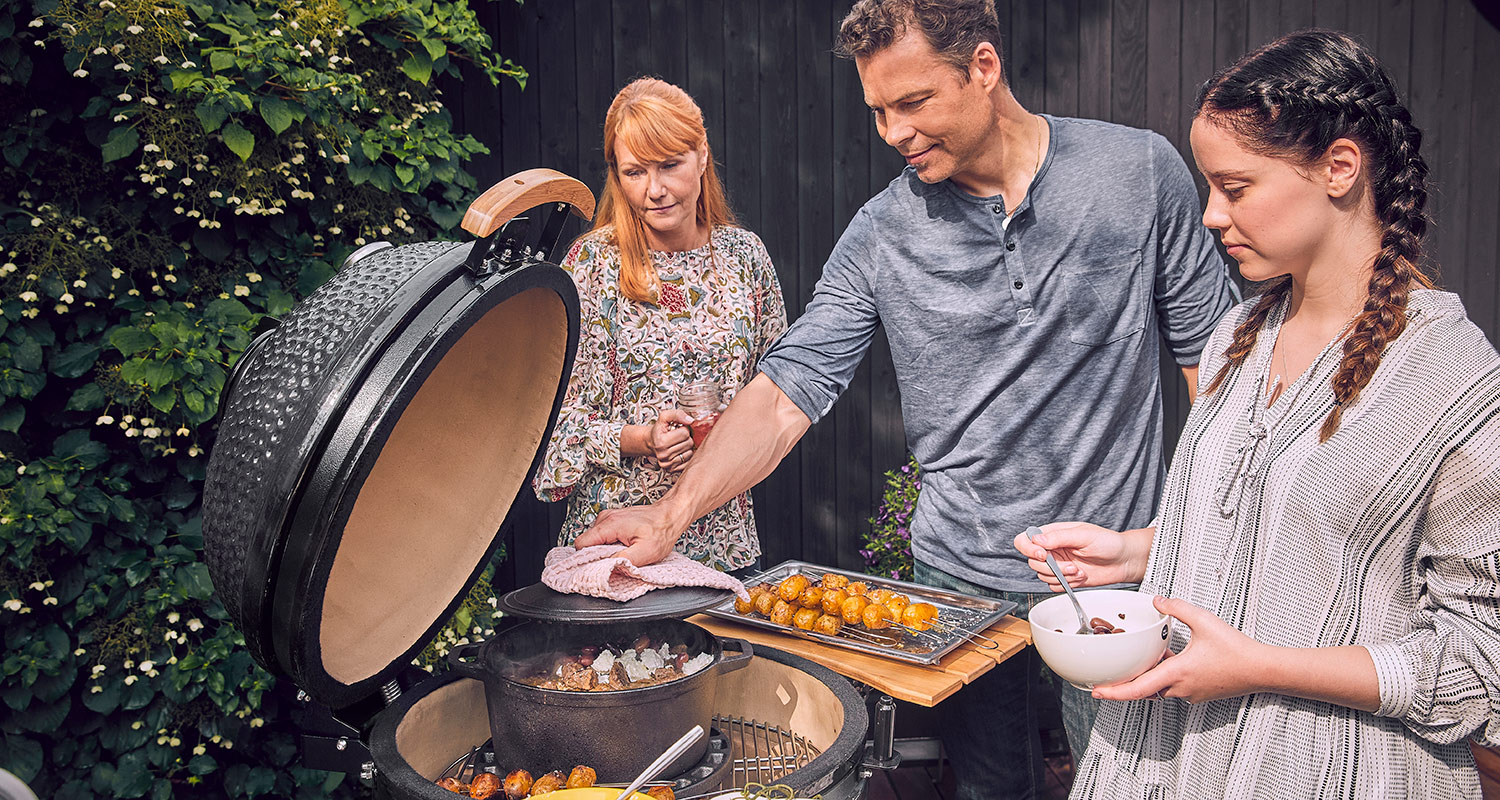 Kolme ihmistä grillaa avatussa mustassa kamado-grillissä, tarjottimella uudet pariloidut perunat.
