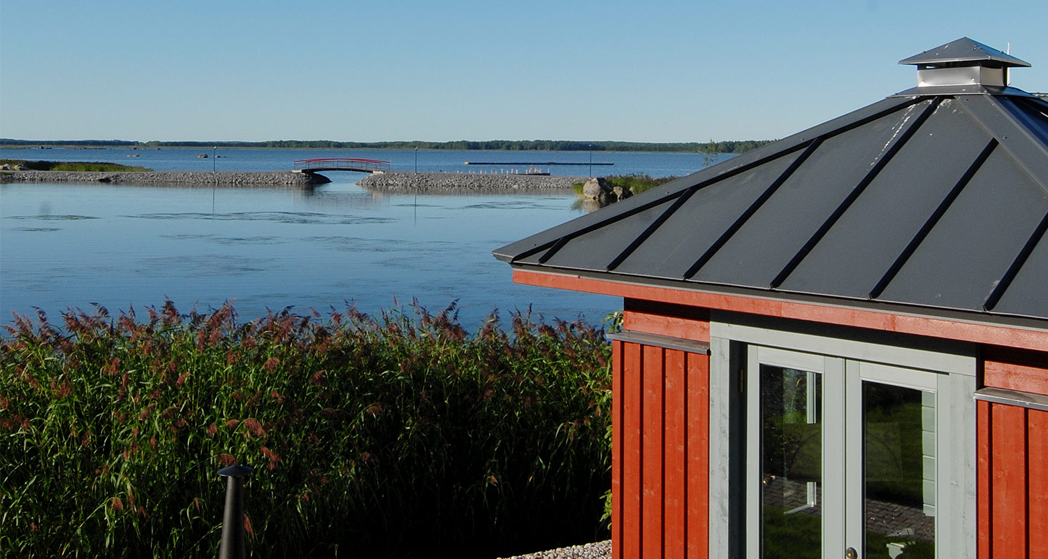 Rannalla punainen paviljonki, etualalla ruskeanvihreää ruohoa, taustalla sininen järvi ja silta.