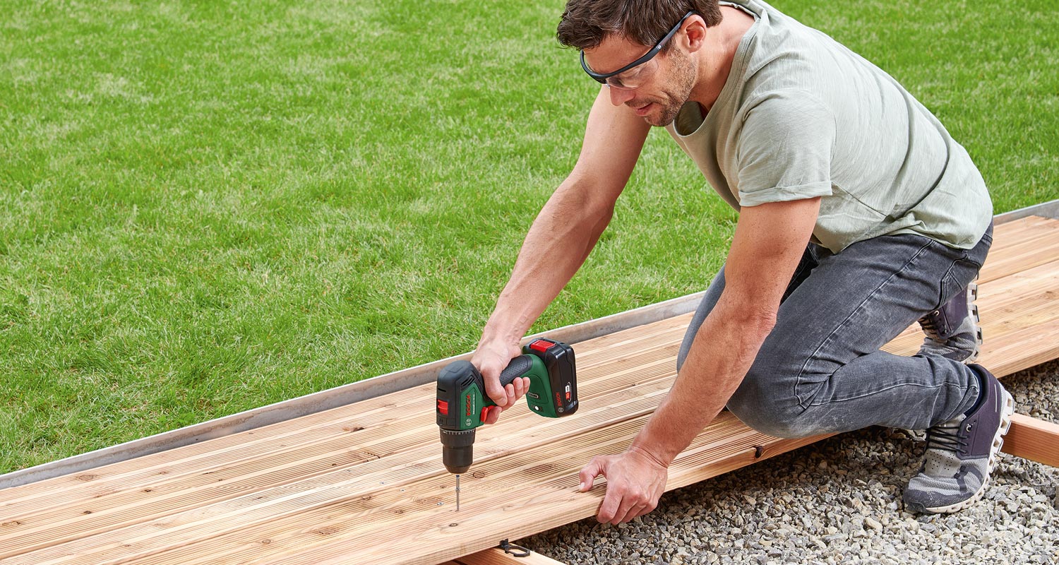 Mies polvillään ruohon laidalla poraa puisia terassilautoja akkuporakoneella.