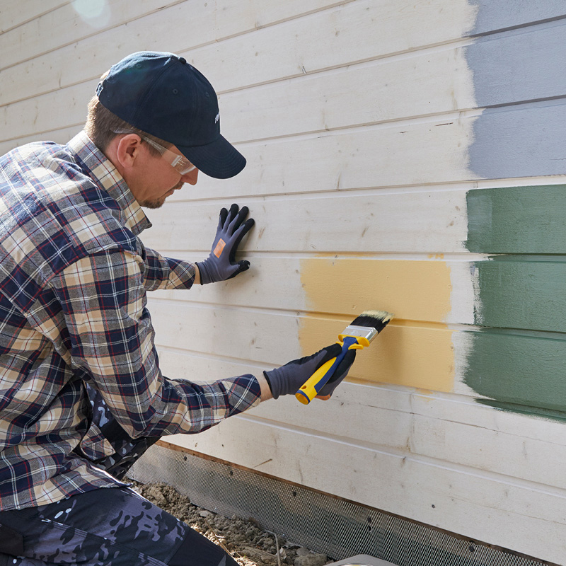 Henkilö maalaa ulkoverhouslautaa keltaisella, vieressä näkyy harmaa ja vihreä värikoelakka.