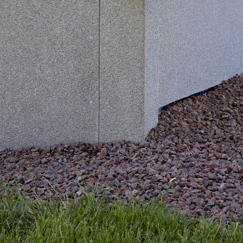 Betoniseinän kulma, sen alla punaruskeaa soraa ja edustalla vihreää nurmikkoa.