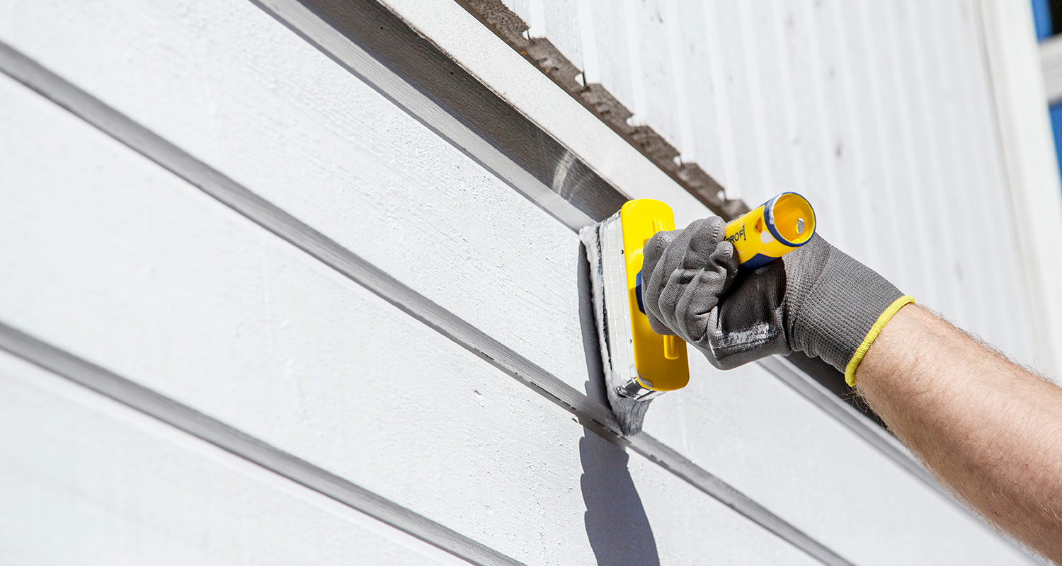 Käsineellä levitetään harmaata tiivistemassaa keltaisella telalla valkoisiin puulautoihin