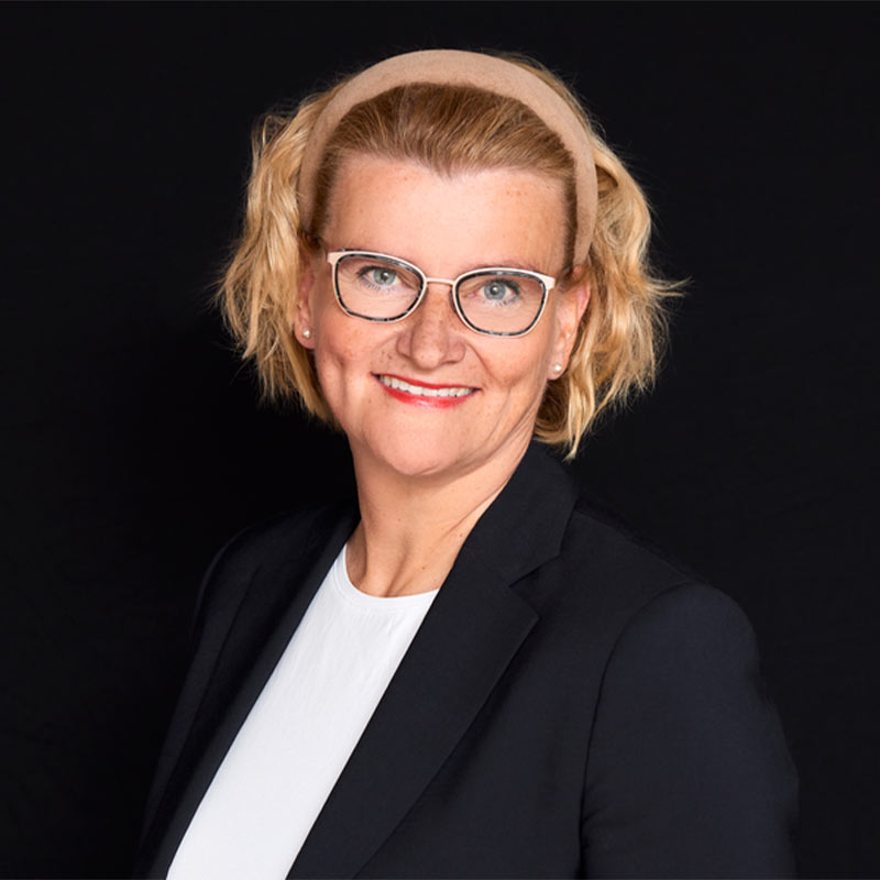 Vaaleahiuksinen henkilö tummalla taustalla mustassa bleiserissä ja valkoisessa paidassa