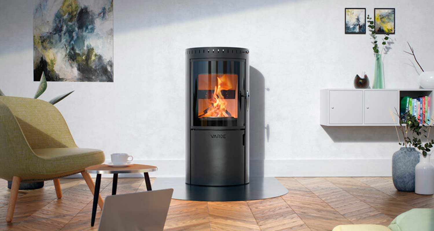 Olohuoneen keskellä musta sylinteritakka palaa, vihreä nojatuoli valkoinen kaappi ja parkettilattia