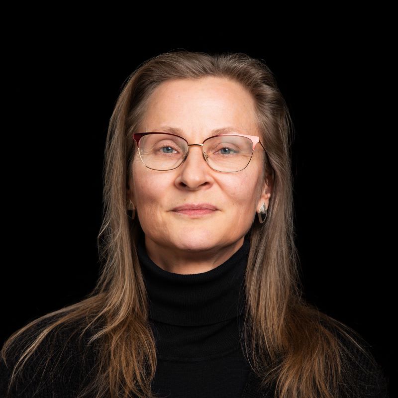 Henkilö, jolla on pitkät vaaleanruskeat hiukset ja musta poolopaita, tummaa taustaa vasten.