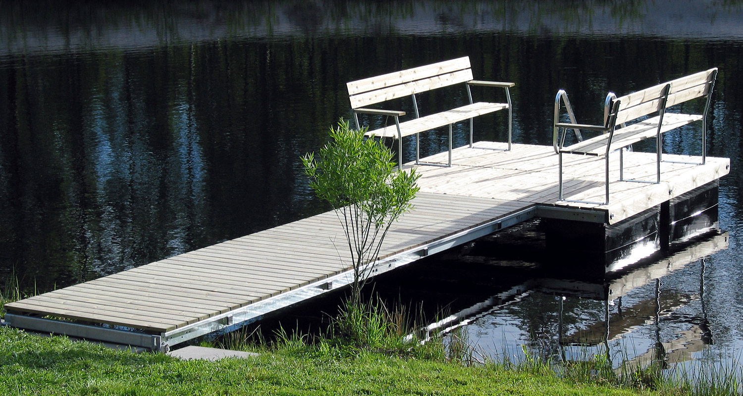 Vaalea puulaituri kahdella penkillä tumman veden äärellä, ruohoa ja pensas rannassa