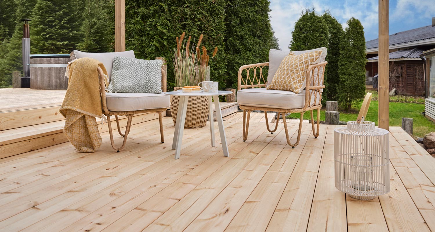 Puinen terassi, rottinkituoleja vaaleine tyynyineen, valkoinen sivupöytä ja kuusiaita.