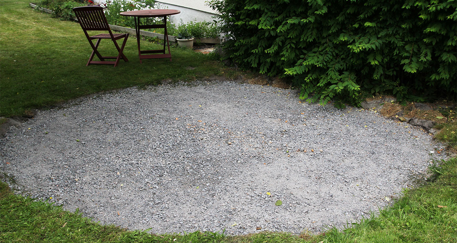 Nurmen keskellä harmaa sorakenttä, takana punaruskea puupöytä ja tuolit, sivulla vihreä pensas.