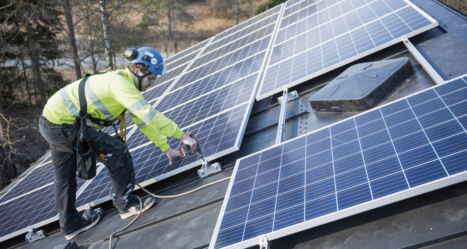 Kattoasentaja kiinnittää sinisiä aurinkopaneeleja keltaisessa takissa metsän reunalla.