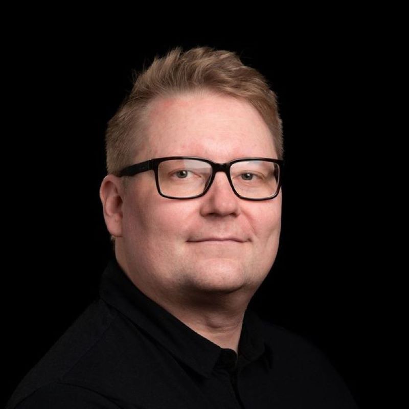 Vaaleahiuksisen henkilön ylävartalokuva mustassa paidassa tumman taustan edessä.