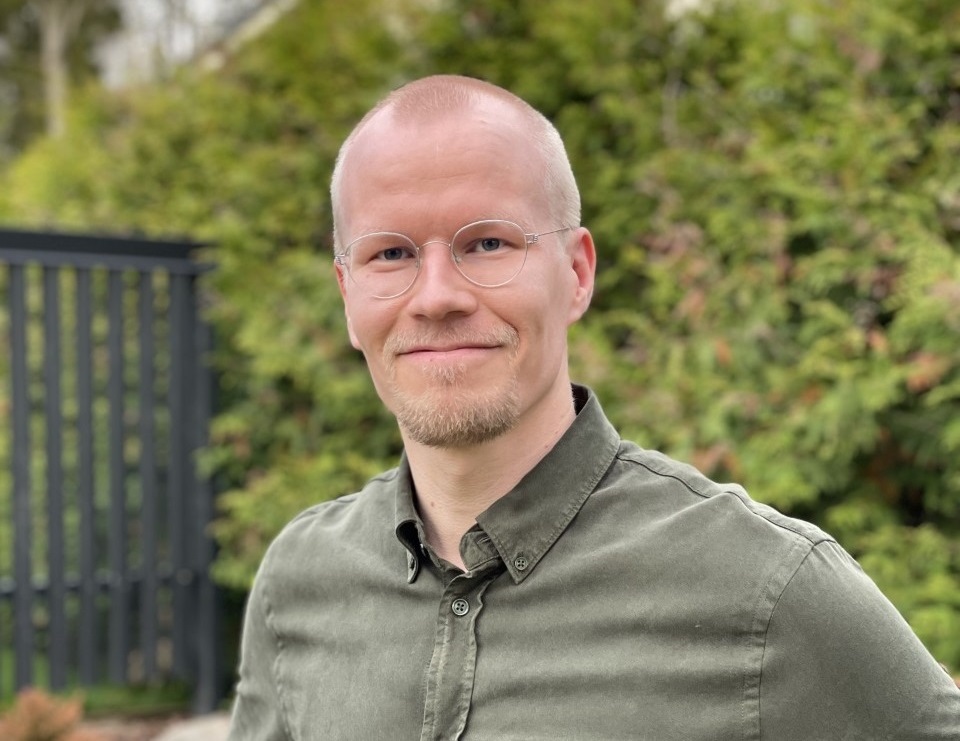 Mies puutarhassa khakinvihreässä paidassa, taustalla musta aita ja pensasaita.