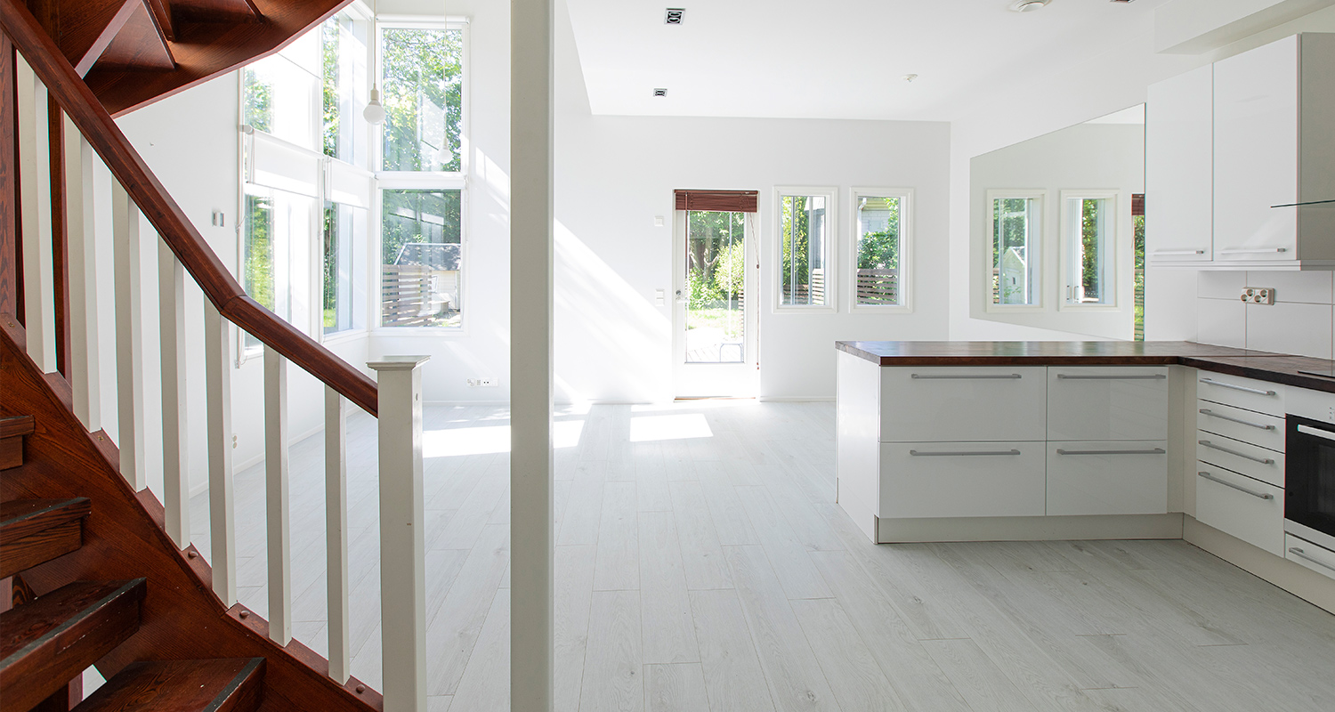 Valoisa olohuone-keittiö: valkoiset seinät, puinen portaat, ikkunaseinä ja keittiösaareke