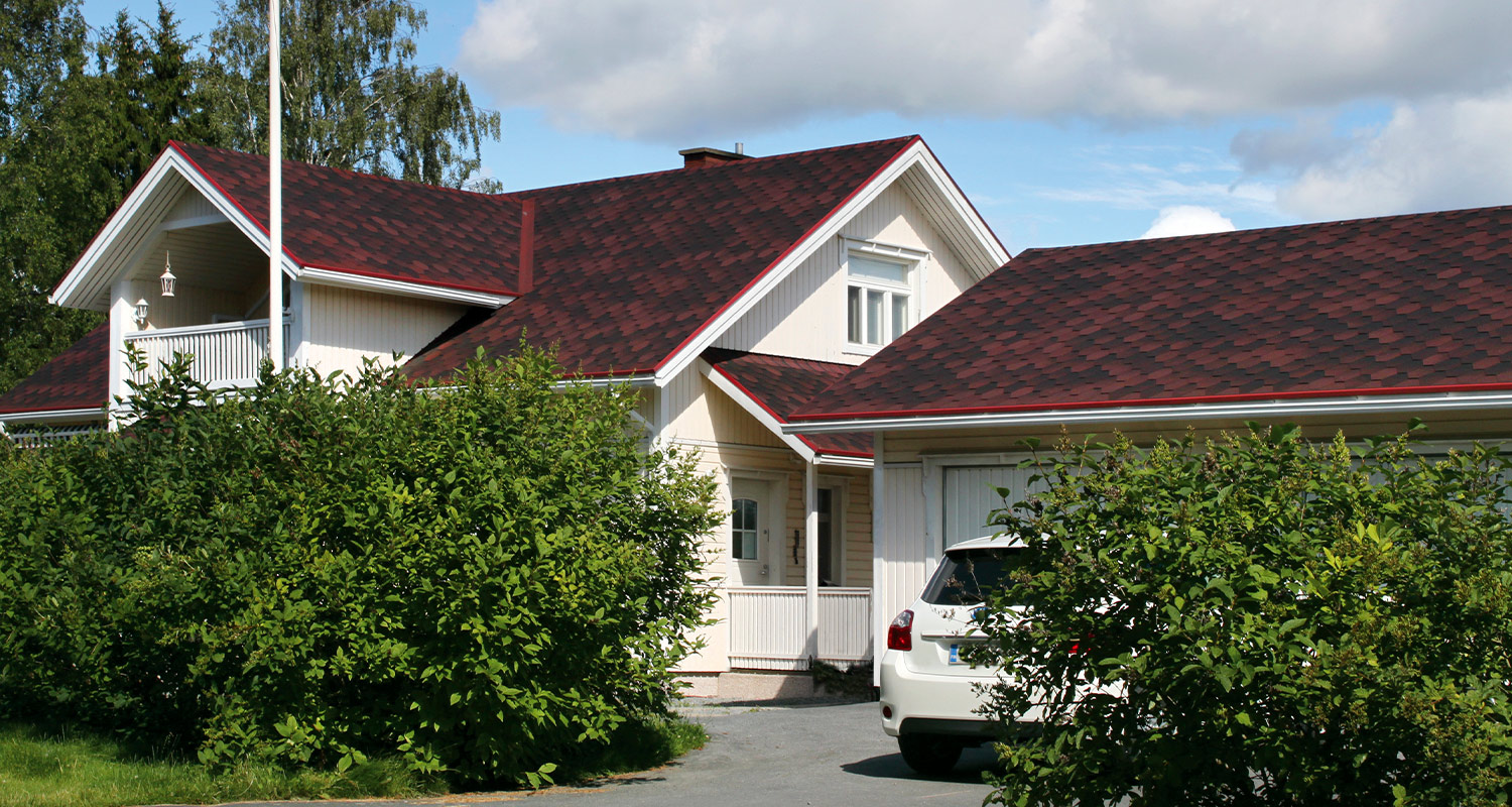 Valkoinen puutalo punaisella katolla, etualalla tiheä vihreä pensas ja valkoinen auto.