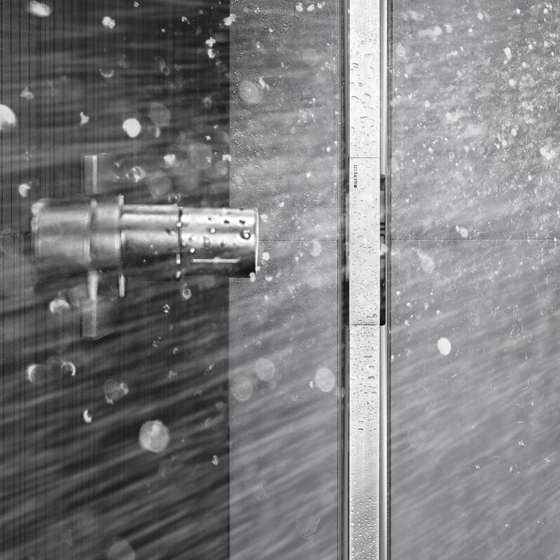 Metallinen suihkupää, lasiseinä ja puunruskeat laatat, suihkuvesi roiskuu.