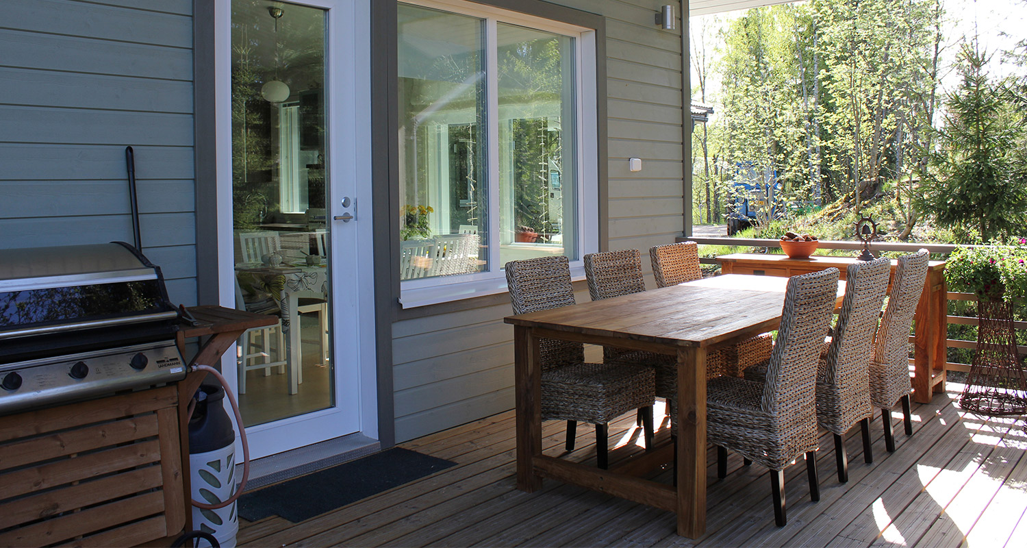 Terassilla puupöytä kuudella punotulla tuolilla, kaasugrilli ja vaalea talon julkisivu.