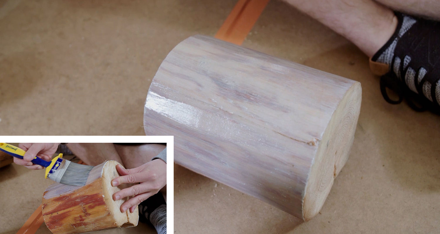 Kädet siveltävät valkoista pohjamaalia puiseen pölkkyyn oranssin remmin päällä.