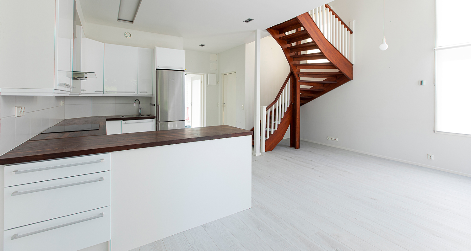 Avoin vaalea keittiö-olohuone, valkoiset kaapit ja tumma puinen kierreportaat.