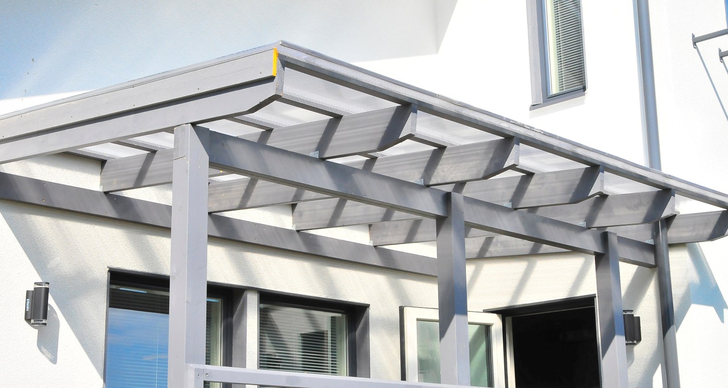 Harmaa pergola valkoisen talon julkisivussa, ikkunat ja avoin ovi varjopiireineen.