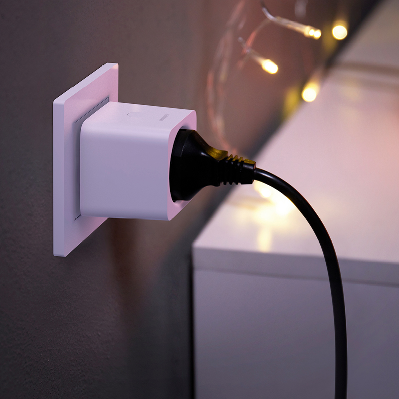 Valkoinen älypistorasia seinässä, musta johto kytketty, taustalla lämpimät LED-valot