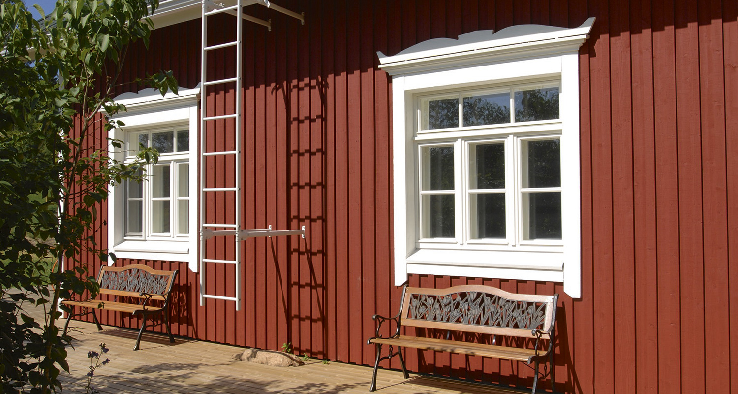 Punainen puutalon julkisivu, valkoiset ikkunat, metallinen tikas ja puiset penkit terassilla