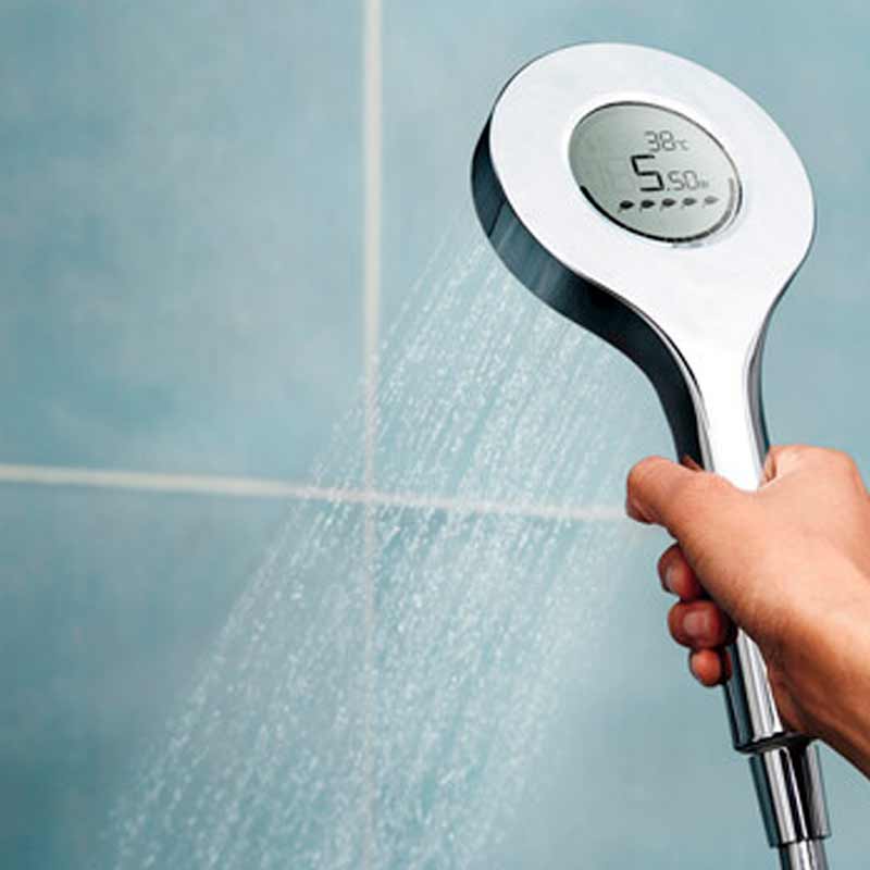 Käsi pitää pyöreää hopeista suihkupäätä diginäytöllä, josta valuu vettä sinistä laattaa vasten.