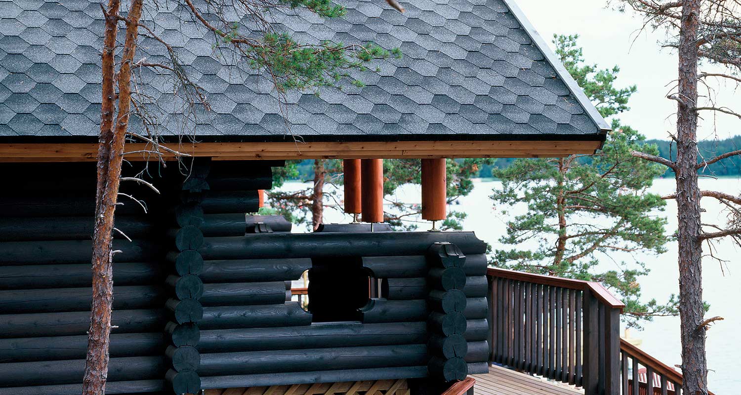 Hirsinen tumma kesämökki vesistön äärellä mäntyjen ympäröimänä