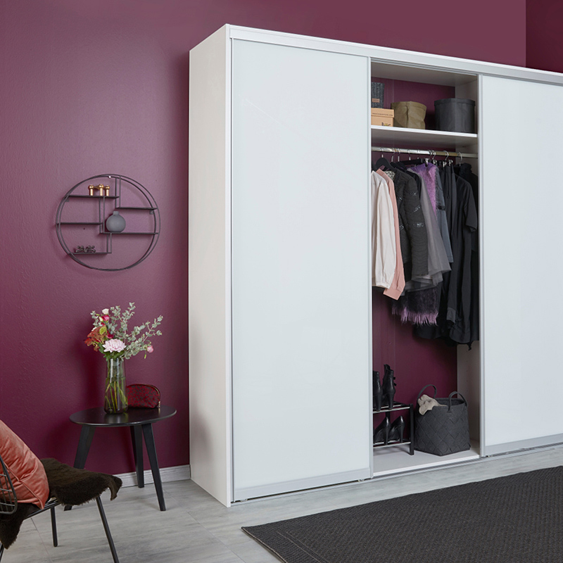 Valkoinen liukuovikaappi viininpunaisella seinällä, avoin keskiosa vaatteita, pöydällä kukkavaasi