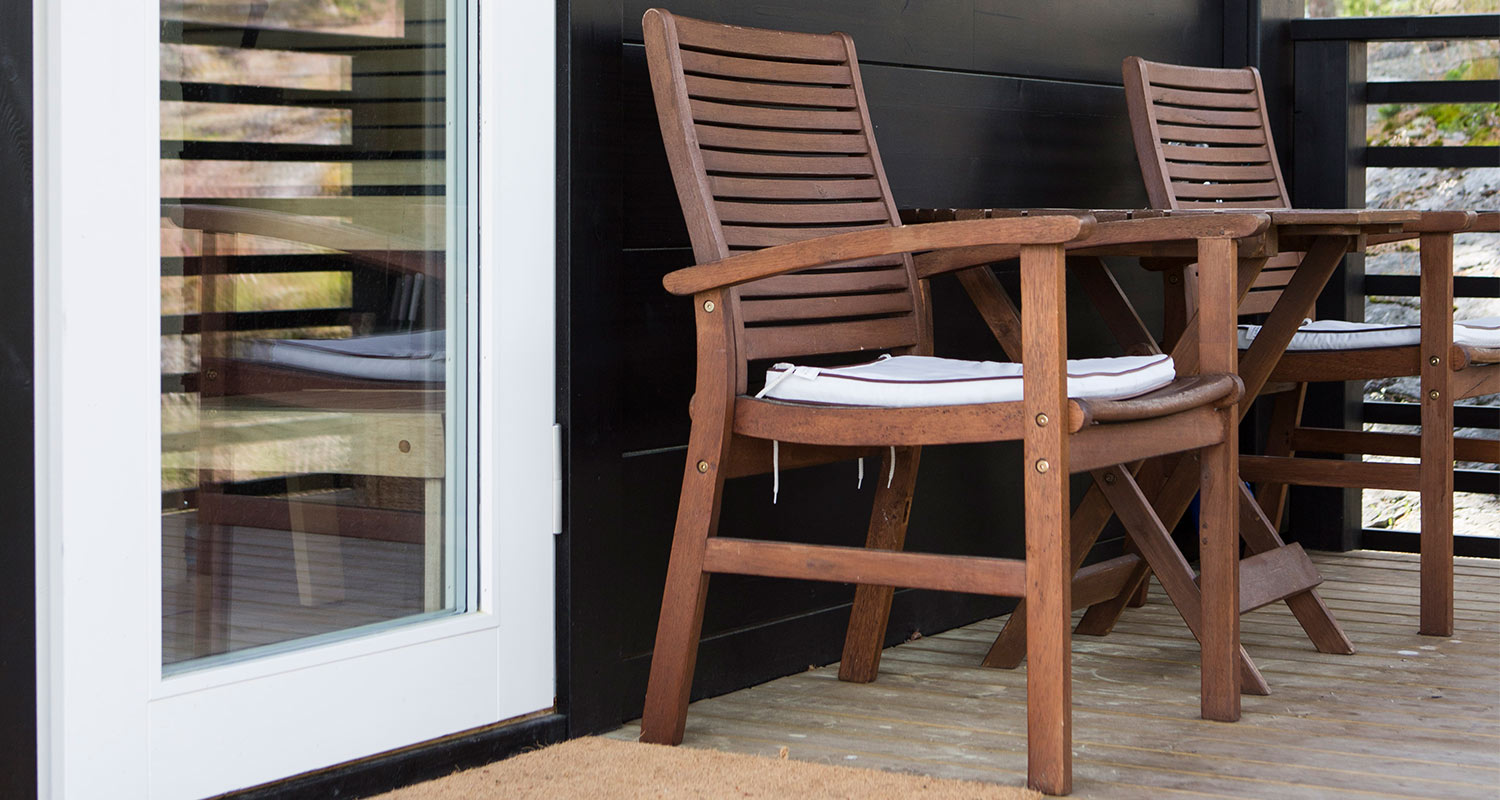 Terassilla kaksi ruskeaa puutuolia valkoisilla istuintyynyillä mustaa seinää vasten.