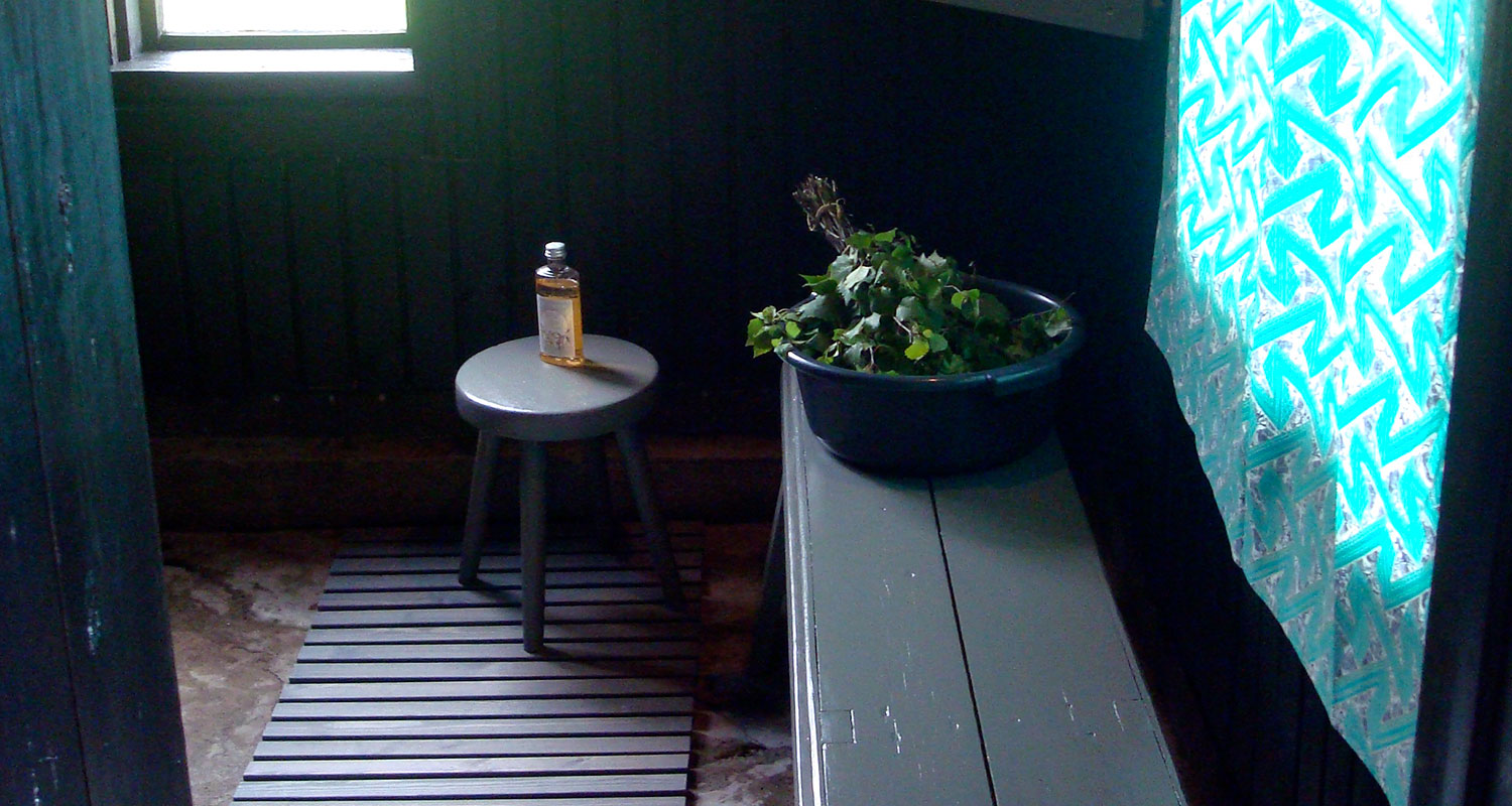 Tummassa saunassa harmaa jakkara öljypullolla, penkillä koivunvihta muoviastiassa ja turkoosi verho.