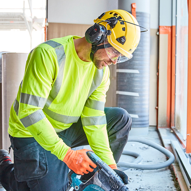 Rakennustyömies neonkeltaisessa paidassa ja keltaisessa kypärässä kyykyssä käyttää sähkötyökalua.