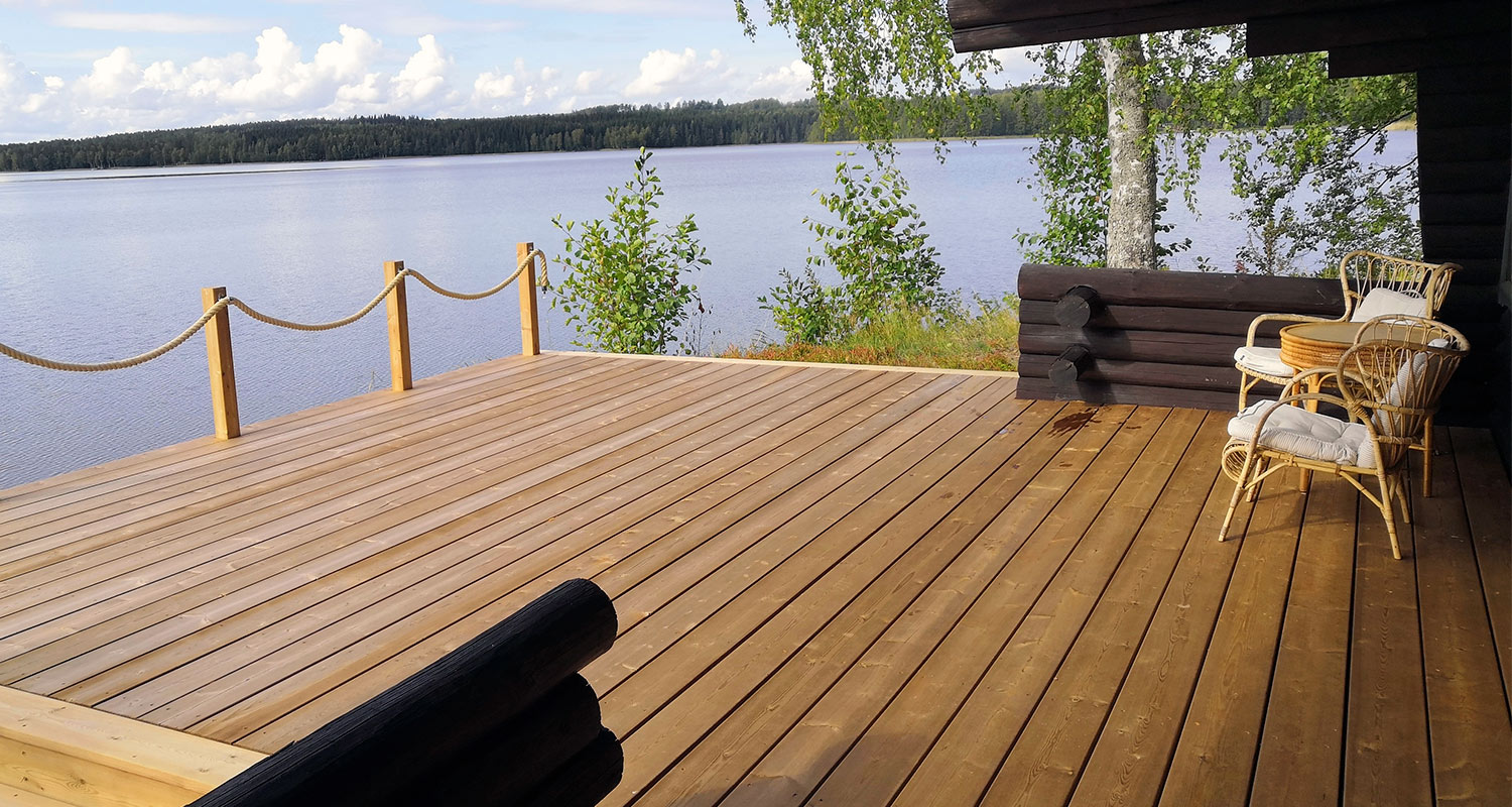Ruskea puuterassi järven rannalla, köysikaide, ratton-tuolit ja pieni pöytä kohti sinistä vesistöä.
