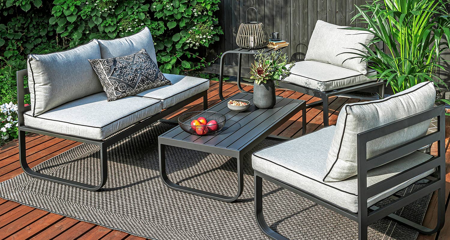 Puulattialla terassin musta metallinen sohvaryhmä harmailla tyynyillä, pöydällä omenoita ja kukkia