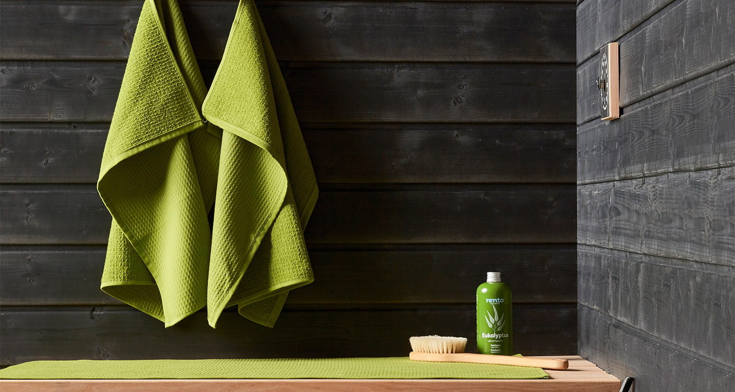 Saunassa musta puupaneeliseinä, limenvihreät pyyhkeet, lauteella harja ja eukalyptuslöylytuoksu