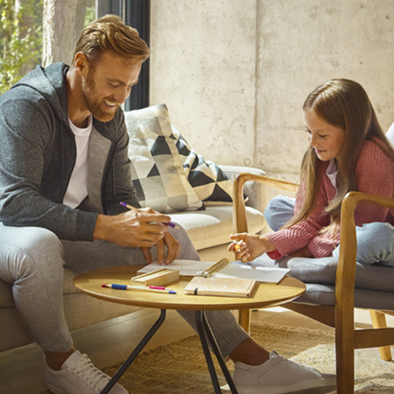 Aikuinen ja lapsi piirtävät läksyjä pyöreän puupöydän äärellä vaaleassa olohuoneessa.