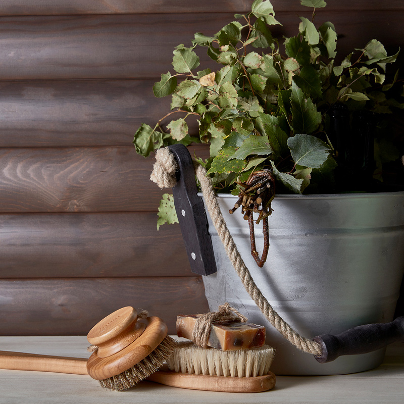 Harmaa ämpäri täynnä koivunoksia, edessä puisia saunaharjoja ja saippuaa.