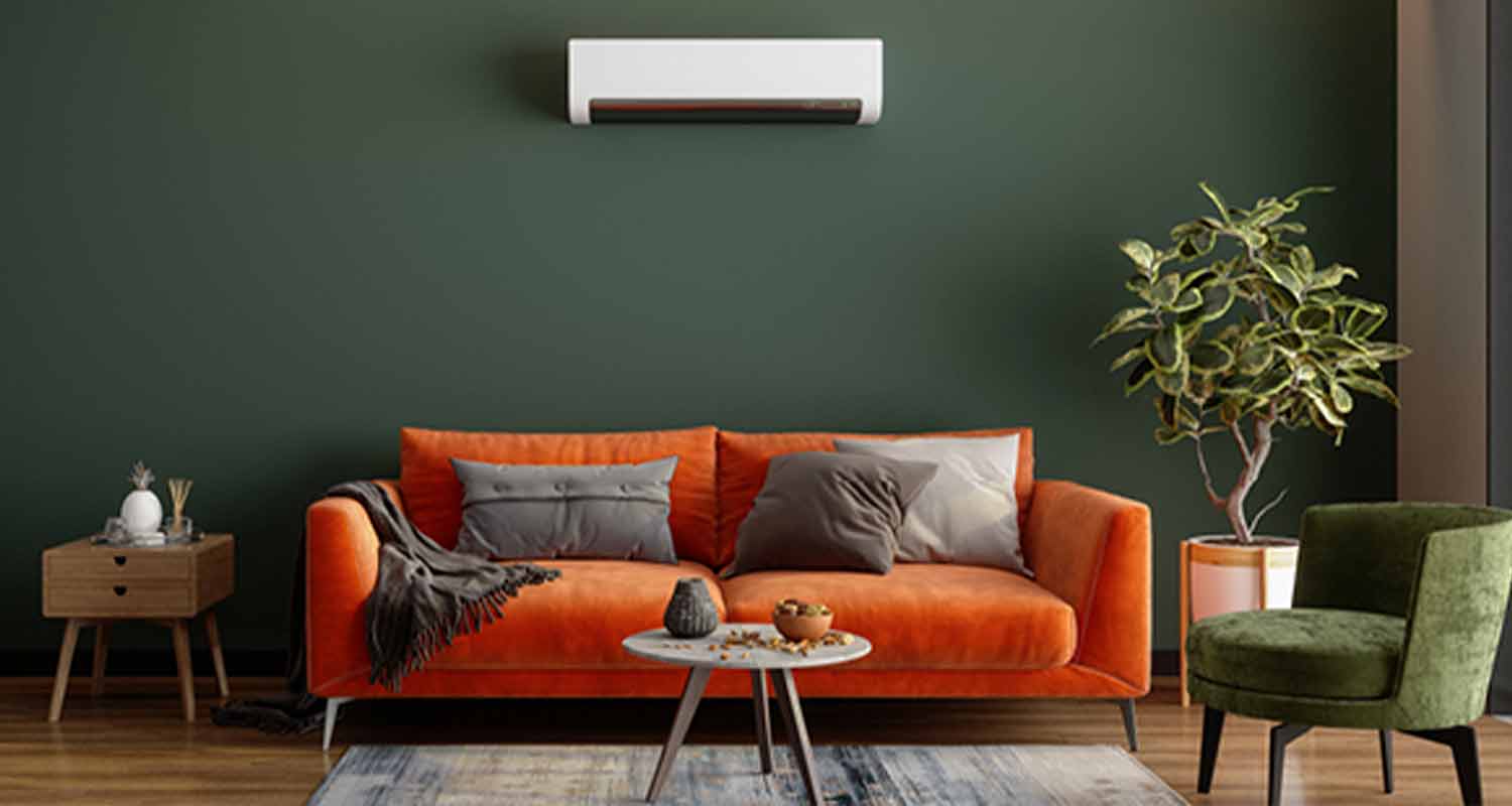 Oranssi sohva tummanvihreää seinää vasten, valkoinen ilmastointilaite, viherkasvi, vihreä nojatuoli.