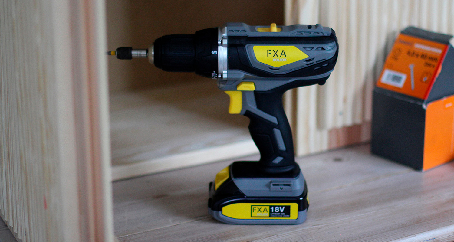 Mustan, harmaan ja keltaisen FXA 18V akkuporakone puupinnalla, vieressä oranssi ruuvilaatikko.