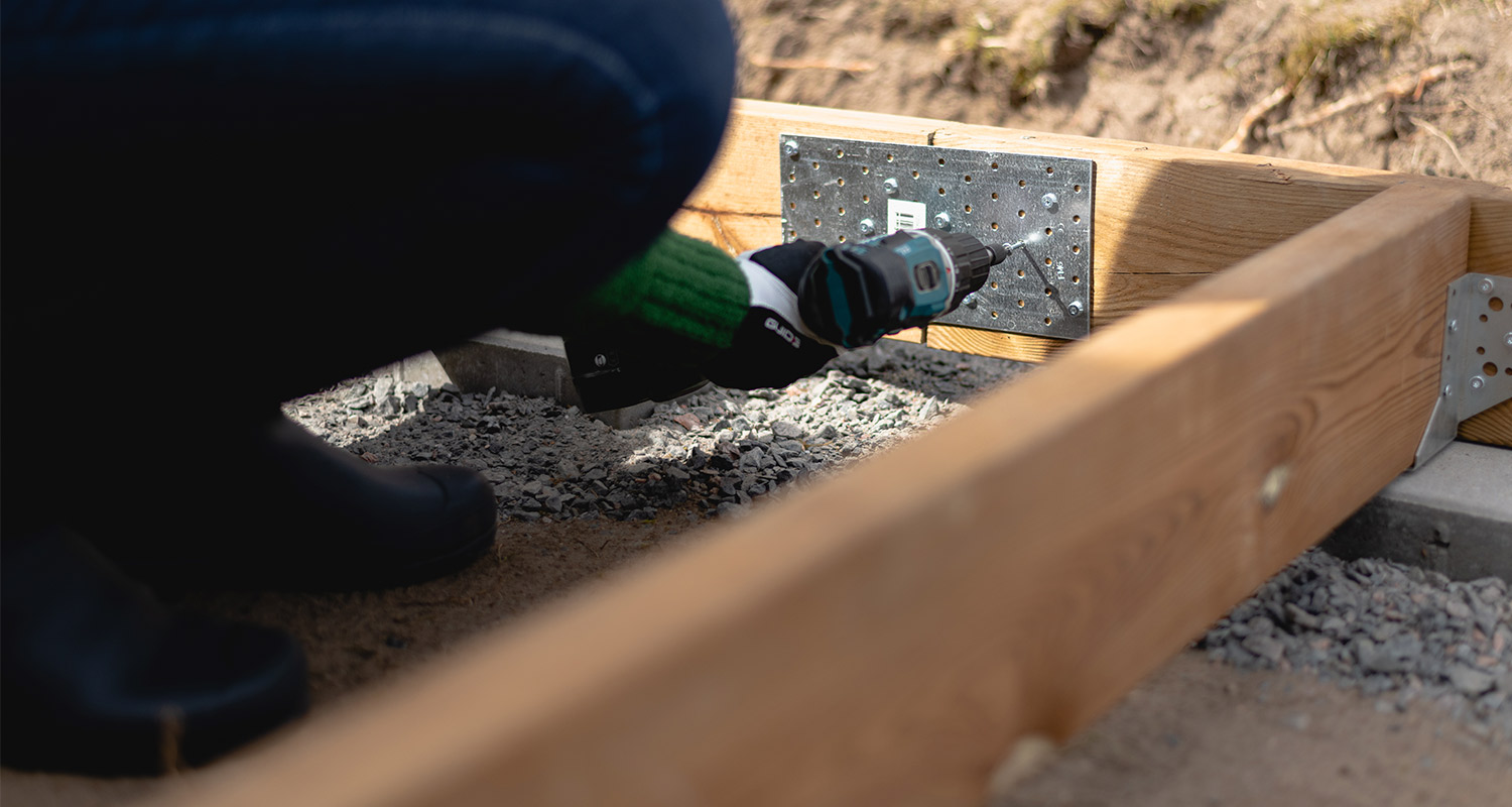 Vihreäkätinen henkilö porakoneella kiinnittää harmaan metallikulman puurunkoon