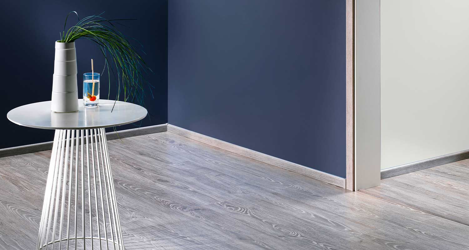 Moderni valkoinen sivupöytä harmaalla puulattialla, tummansininen seinä, maljakko ja juomalasi.