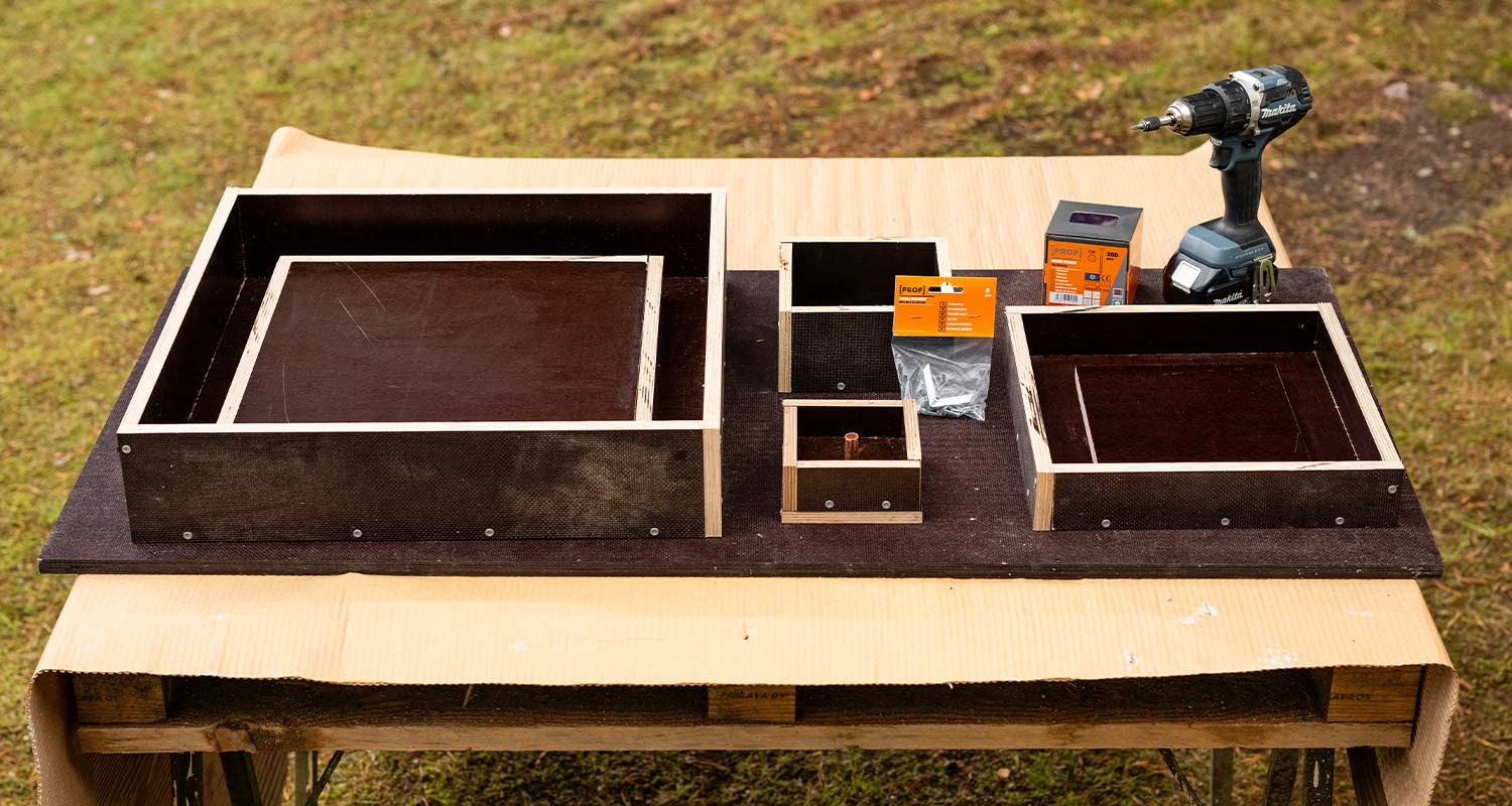 Ulkopöydällä eri kokoisia musta–vaaleapuisia laatikkokehikoita, ruuvipakkauksia ja porakone.